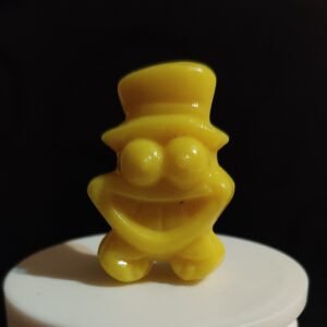 Figura Gogo Crazy Bones amarilla con sombrero de copa y gran sonrisa, colocada sobre base blanca y fondo negro